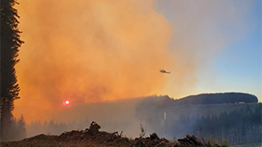 Image of smoke from the Lane 1 fire in Oregon. The sun is reflecting against the smoke, making the sky on the left side of the image appear very orange. A helicopter can be seen on the right side of the image.