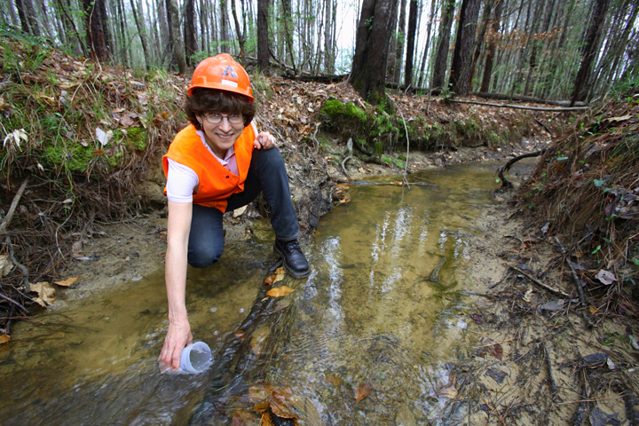 A young intern takes a water sample from a creek while wearing a hardhat and safety orange vest.