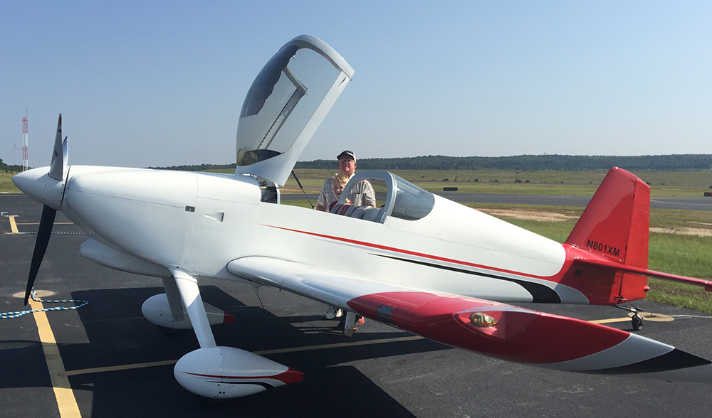 Image of Don Moore and his grandson outside of Don's airplane. The plane is white with red wing tips and tail and has a propeller on the front of the plane. The plane's canopy is open. Don can be seen holding his grandson on the far side of the plane.