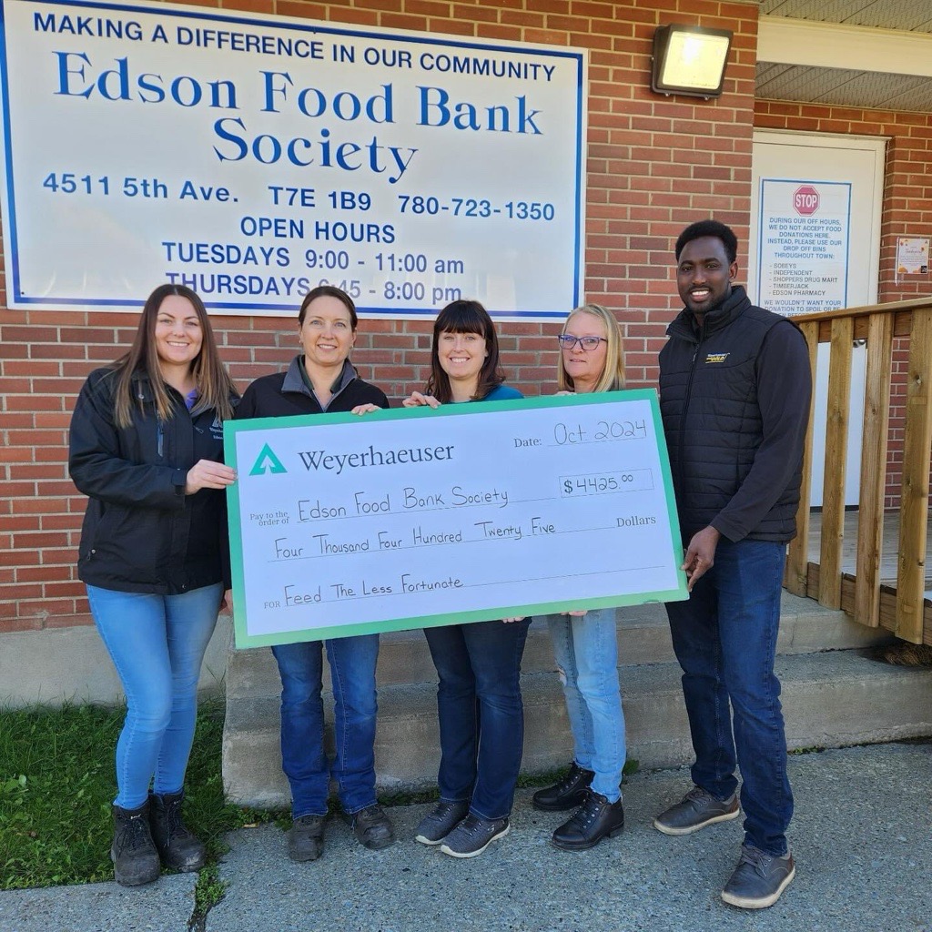 Image of Abdul and colleagues presenting a check to the Edson Food Bank. Edson is standing to the right with four other women holding a large check in front of a sign for the Edson Food Bank Society.