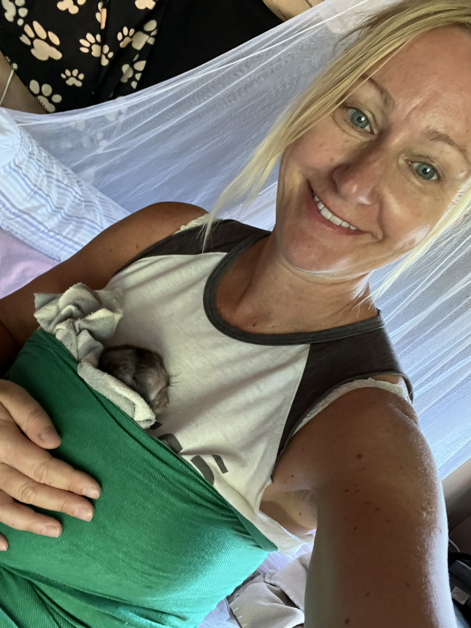 Image of Lana holding a baby vervet monkey. The vervet is in a sling and its face is against Lana's shirt. The harness is green.