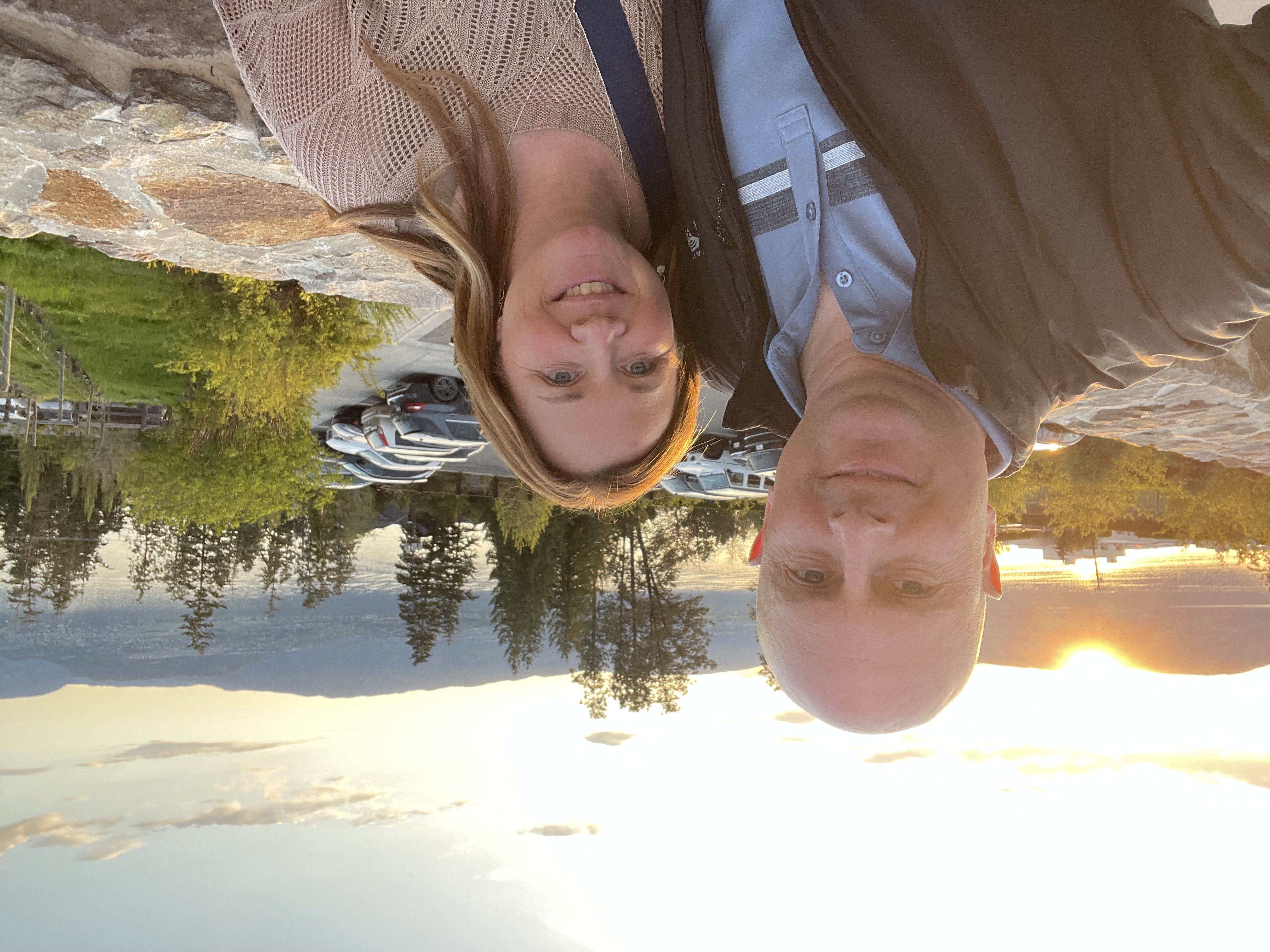 Image of Kerri and her husband, Jeff, at a local winery. They are standing with the sun setting behind them over a mountain. In front of the mountain is a lake, with trees lining the lake.