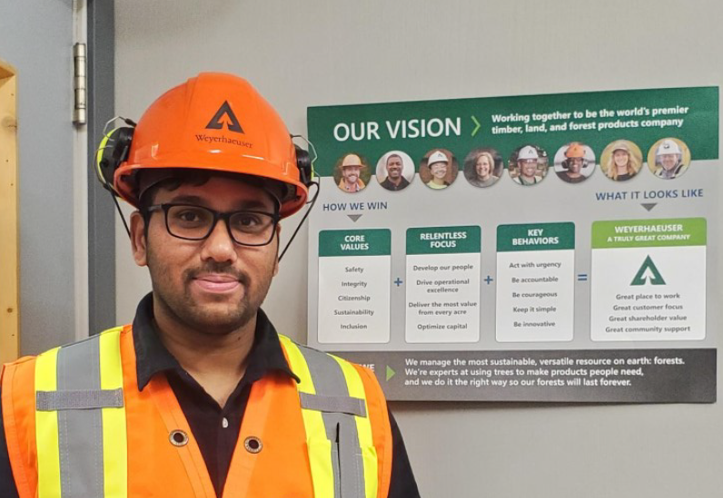 Image of Sathish Nachimuthu standing next to a sign that displays our values as a company. Sathish is wearing an orange safety vest and orange hard hat.