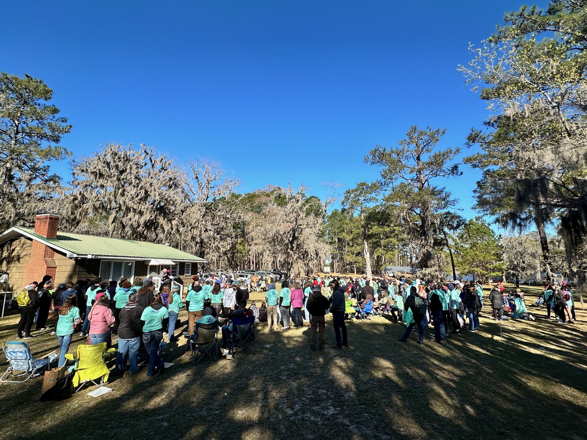 Image of the some of the nearly 500 visitors who came to Cool Springs for the 2024 Envirothon. The visitors are dressed in an array of colors and are standing in a field in front of numerous trees.