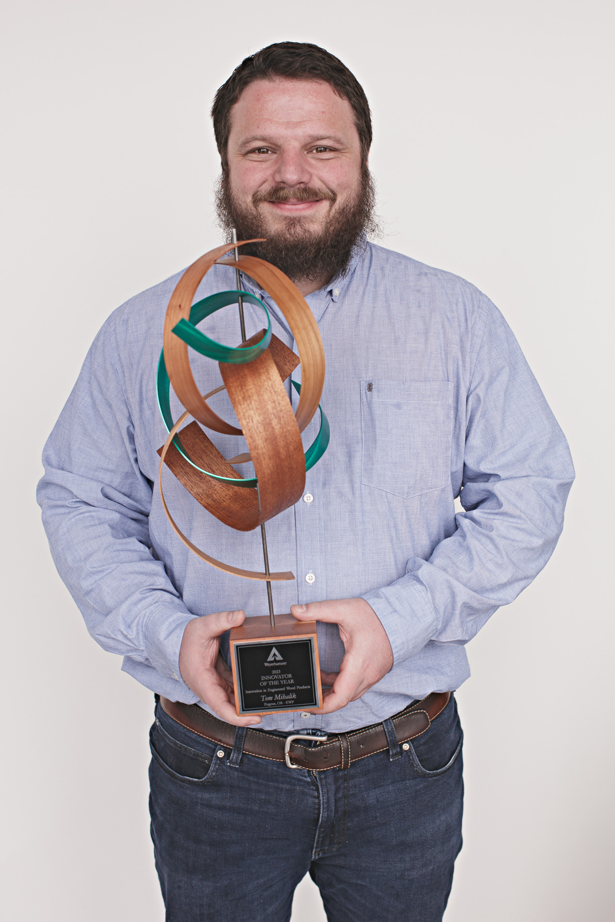 Tom stands against a white backdrop while holding the 2023 Innovator of the Year award, which is a sculpture of wood grain and green loops with a metal base
