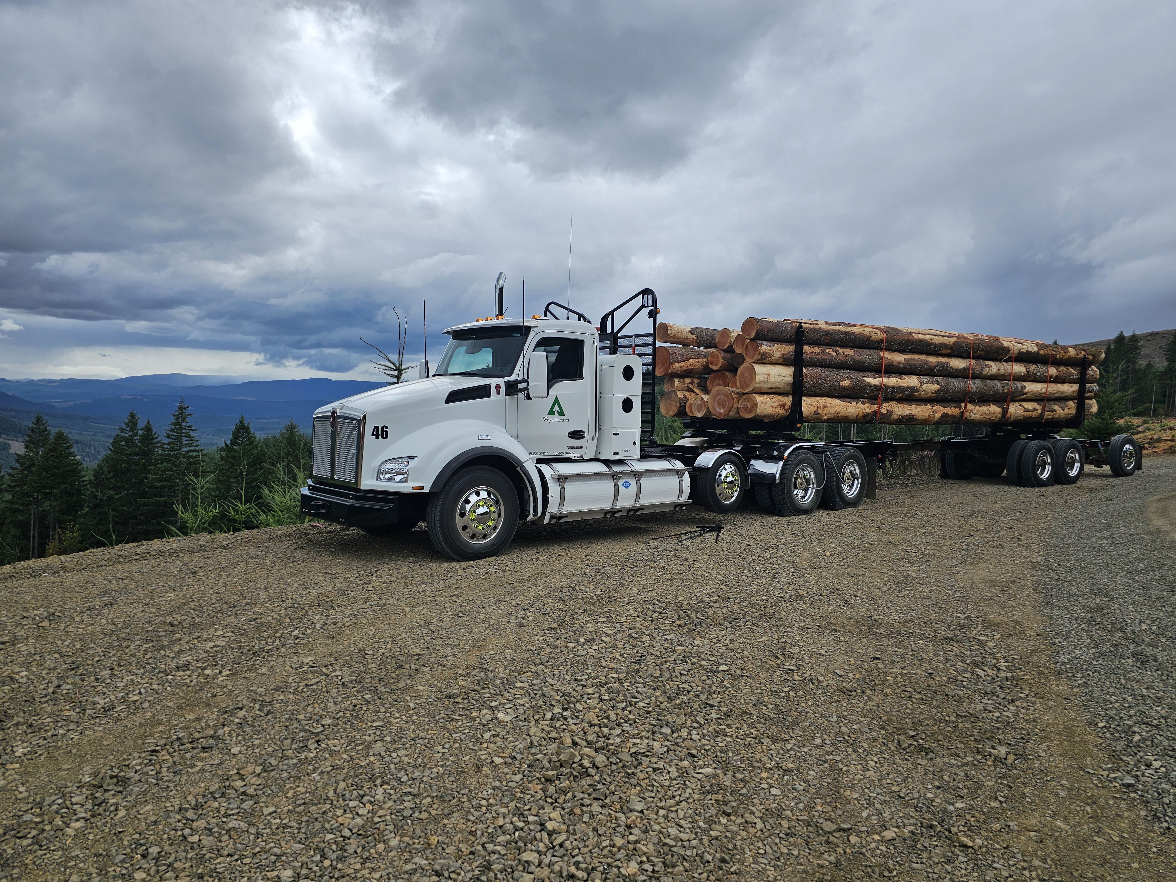 Image of a natural gas-powered log truck. The truck's cab is white and it is on a patch of gravel that overlooks a treeline. The sky above is very cloudy and mountains can be seen in the background.