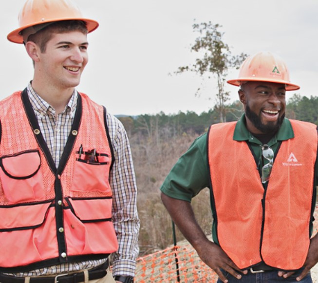 Two individuals wearing orange safety vests and hardhats smiling as they stand in the outdoors.