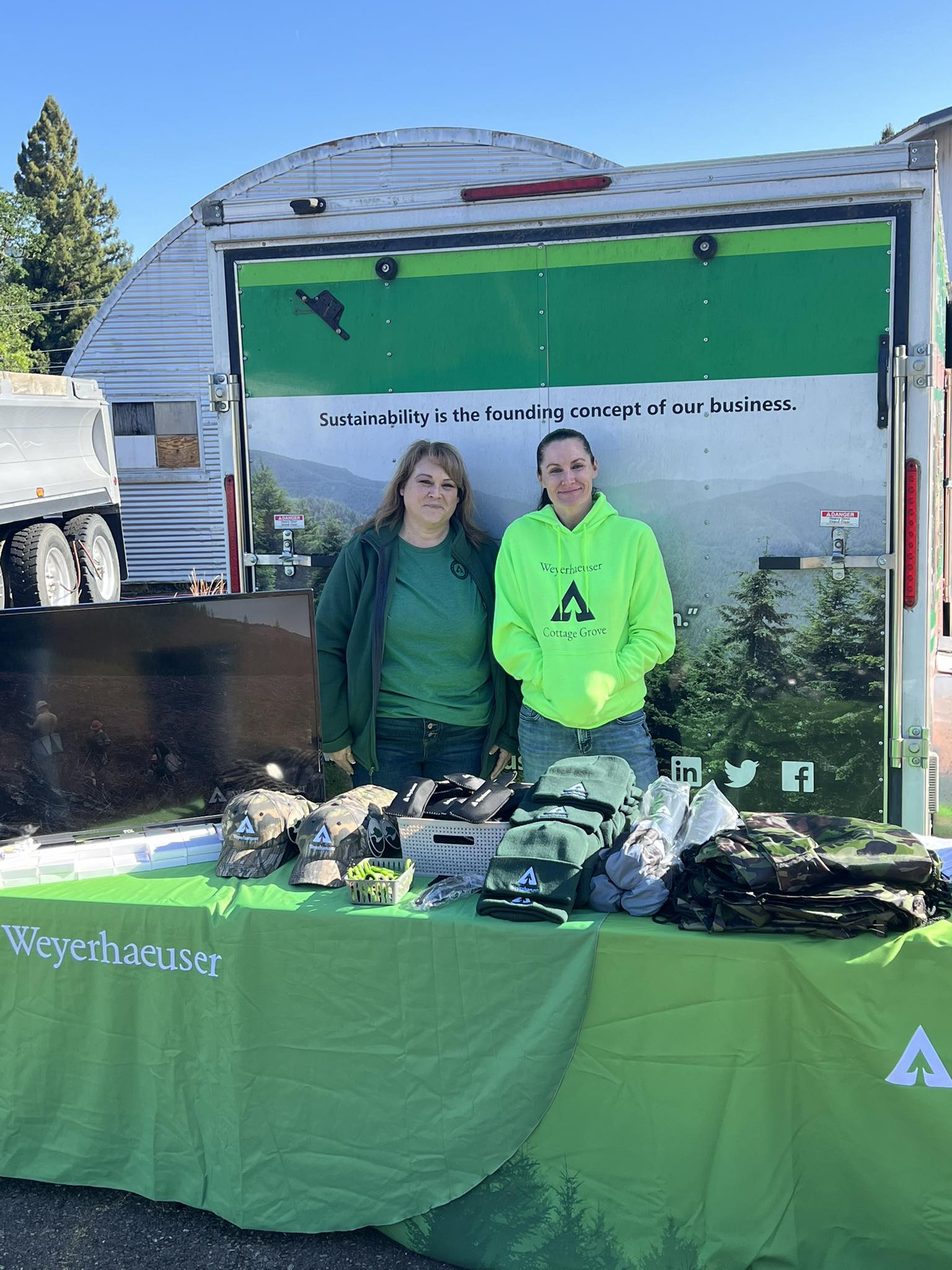 Image of Amber, right, and Tracy Ollivant, lumber human resources business partner, preparing for a local job recruitment fair. Amber wears a fluroscent green sweatshirt and stands behind a table that is covered with a green Weyerhaeuser-themed tablecloth