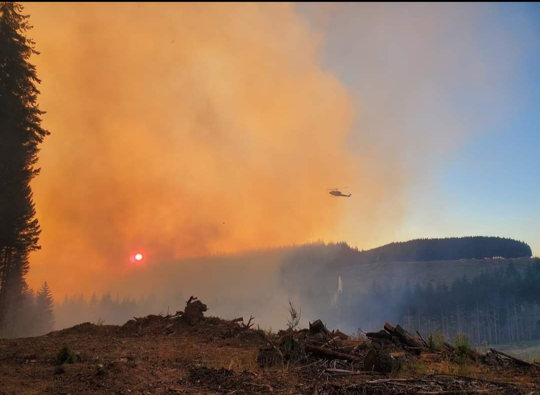 Image of smoke from the Lane 1 fire in Oregon. The sun is reflecting against the smoke, making the sky on the left side of the image appear very orange. A helicopter can be seen on the right side of the image.