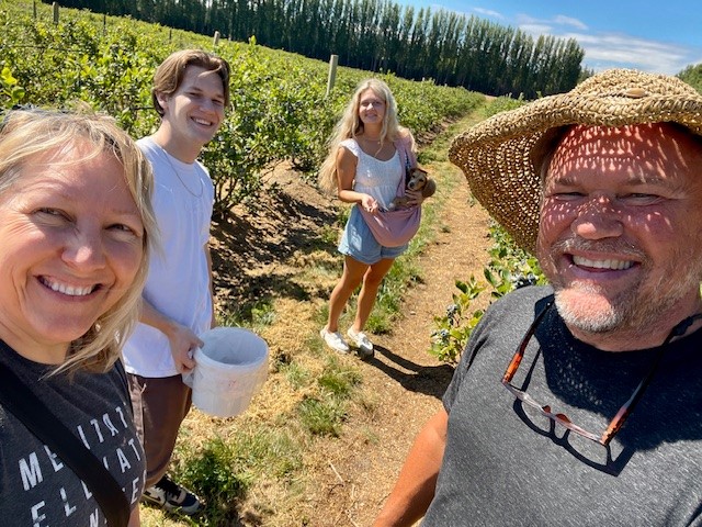Image of Steve and his family picking berries. Steve is on the right side of the picture and is wearing a straw hat that is casting shade on his face. His wife is on the left side and his two children are in the middle. Fields of berries can be seen.