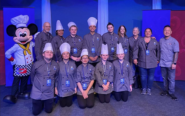 Image of Deborah with the Enterprise Culinary Team at a cooking competition at Disney World. The chefs are all wearing matching coats, an some wear toques on their heads. Along with the team are Mickey Mouse and celebrity chef Jet Tila.