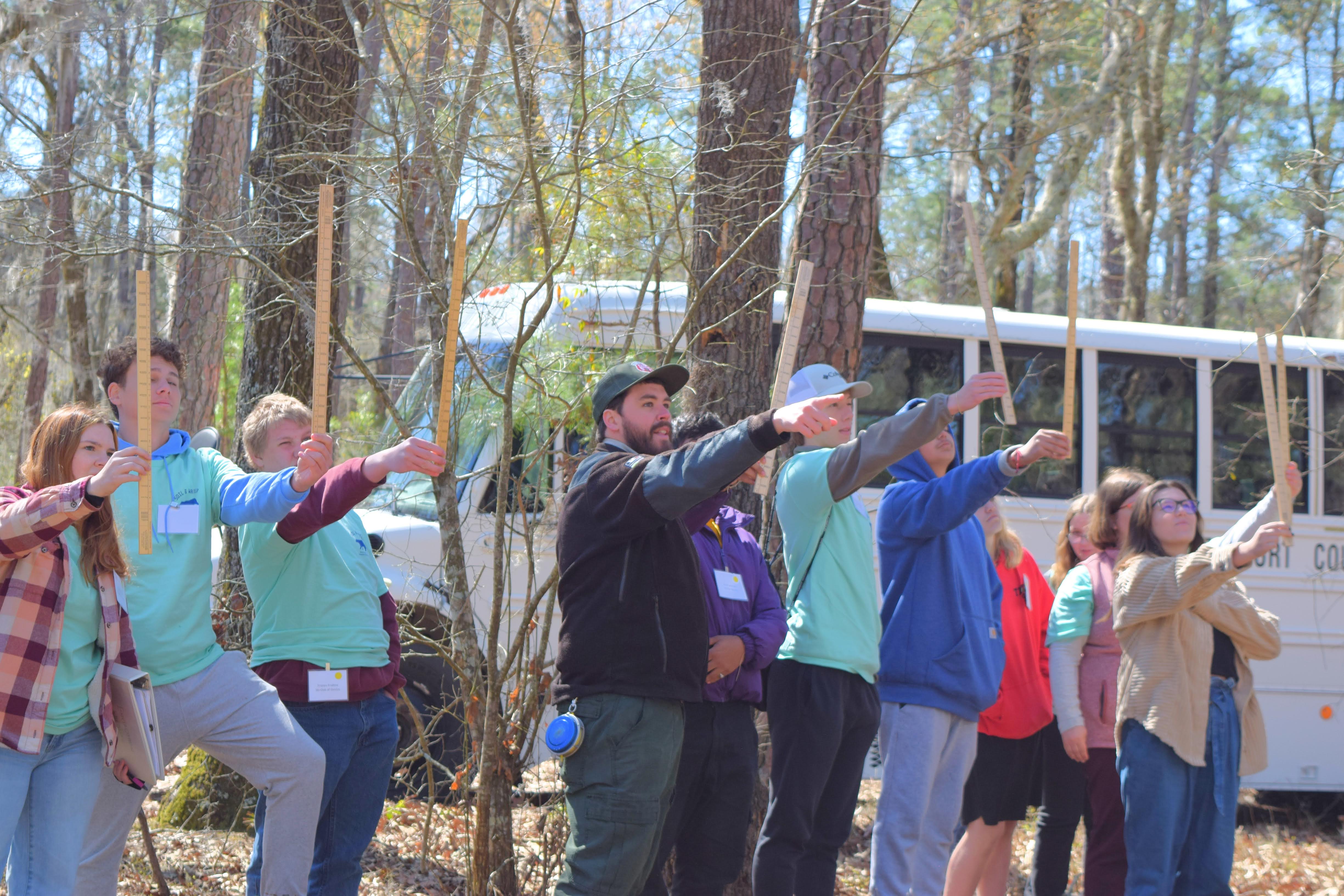 Image of people taking part in this year's Envirothon. A group of approximately 11 people wearing a variety of shirts, some turquoise, others a variety of colors, hold sticks in the air.