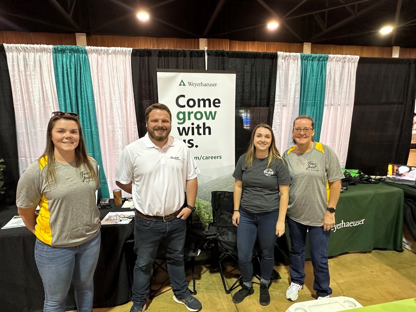 Tom and three other people stand in a row in front of a curtained booth at a recruiting fair with a banner reading "Come Grow With Us" in the background.
