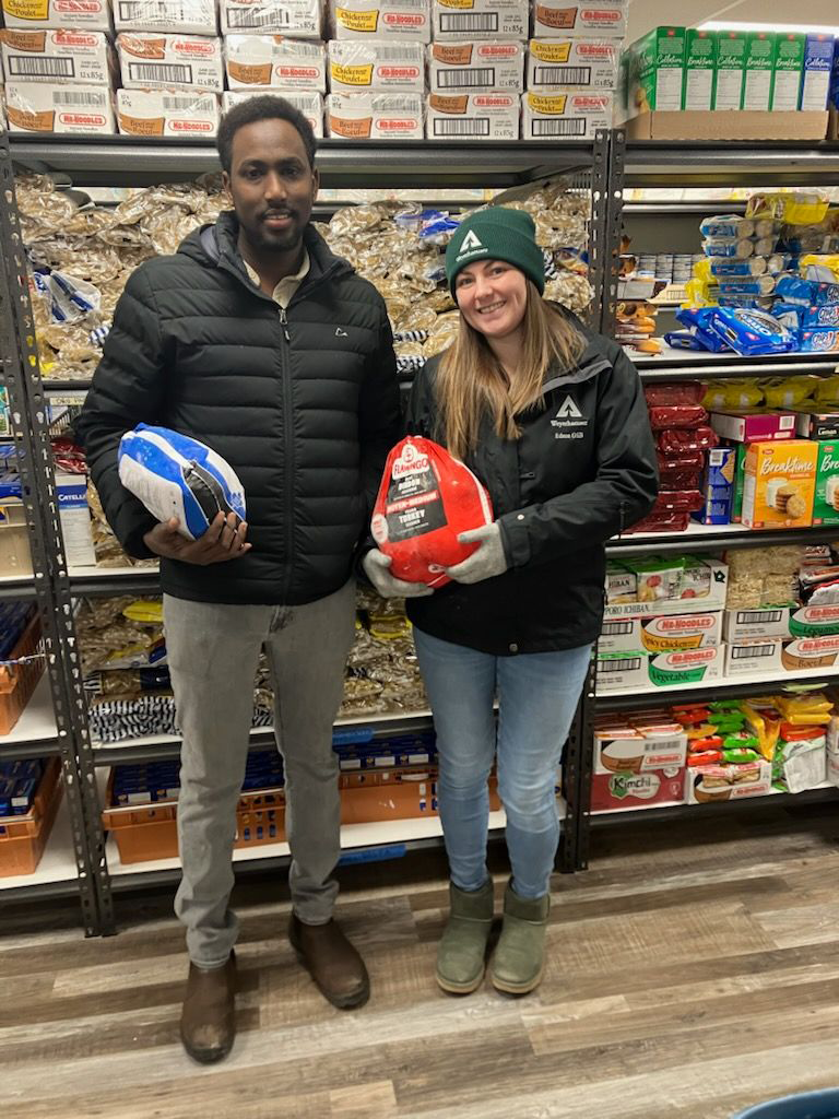 Image of Abdul and his colleague Jasmine Hagen holding turkeys in the food bank. Adul is standing to the left and Jasmine to the right. They are each wearing coats. Abdul's turkey is in a white and blue package, while Jasmine's is in a red package.