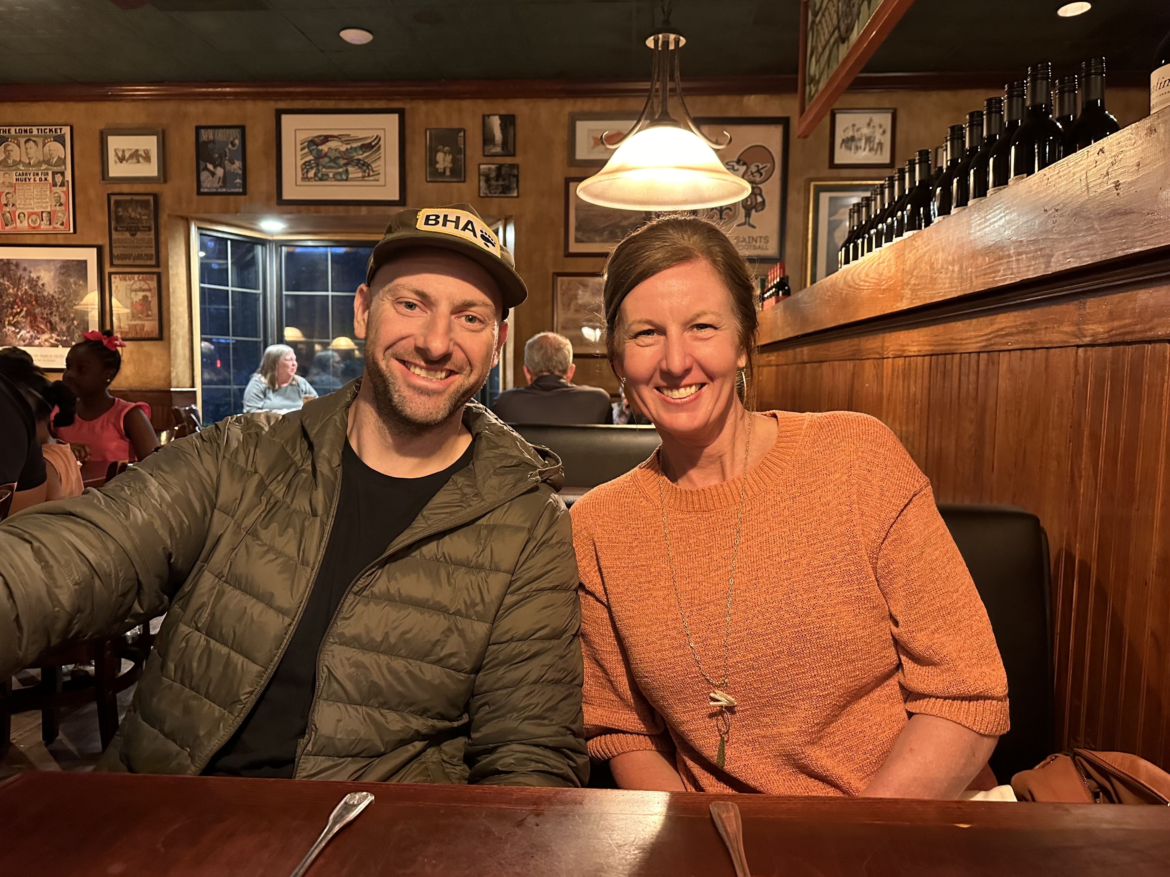 Image of Christie, sitting on the right in an orange sweater, and Michael Kelly, who is wearing a baseball cap and a jacket. They are sitting at a table in a restaurant.