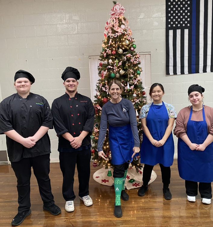 Image of five people standing in front of a Christmas tree. Deborah is standing in the center and is pointing at her green Weyerhaeuser socks.