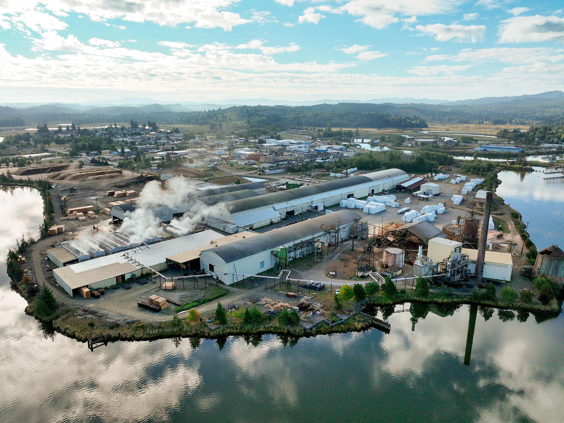 Image of an aerial vieew of the Raymond mill taken by a drone. The mill is surrounded on three sides by water, and forests and mountains can be seen in the background.