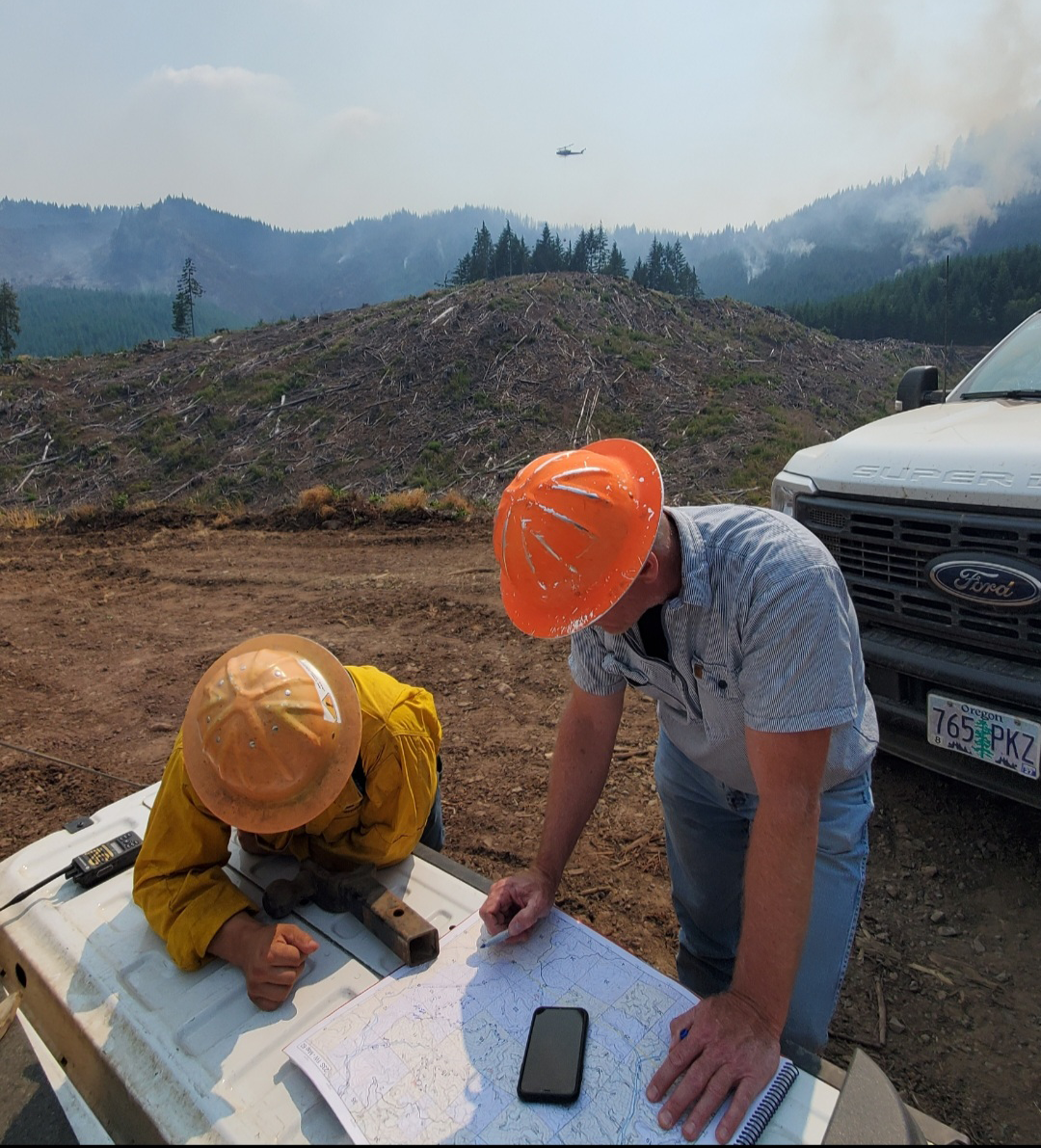 Image of two men wearing hardhats and looking at maps as they are coordinating ways to fight the Lane 1 fire. A helicopter can be seen in the sky on the background and smoke is rising on the upper right side of the image. 