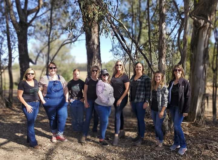 Image of nine women wearing jeans and shirts, with some wearing flannel shirts or jackets overtop. They are standing in front of a group of trees.