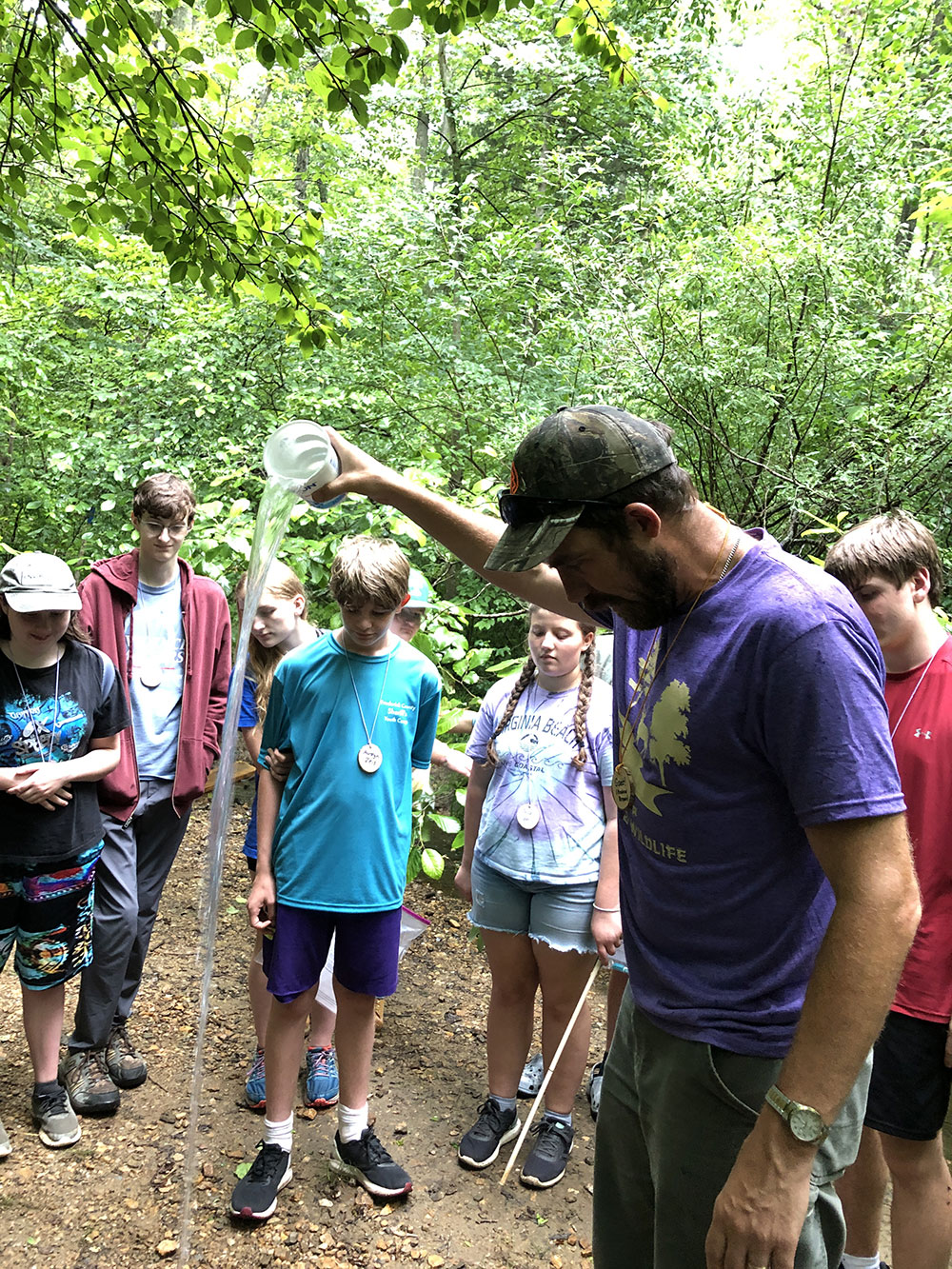 Image of Andrew performing a demonstration to a group of kids. He's wearing a purple t-shirt and is holidng a cup of water above shoulder height. The water is pouring down ionto the ground. Kids are surrounding Andrew and are all looking down at the water landing on the ground.