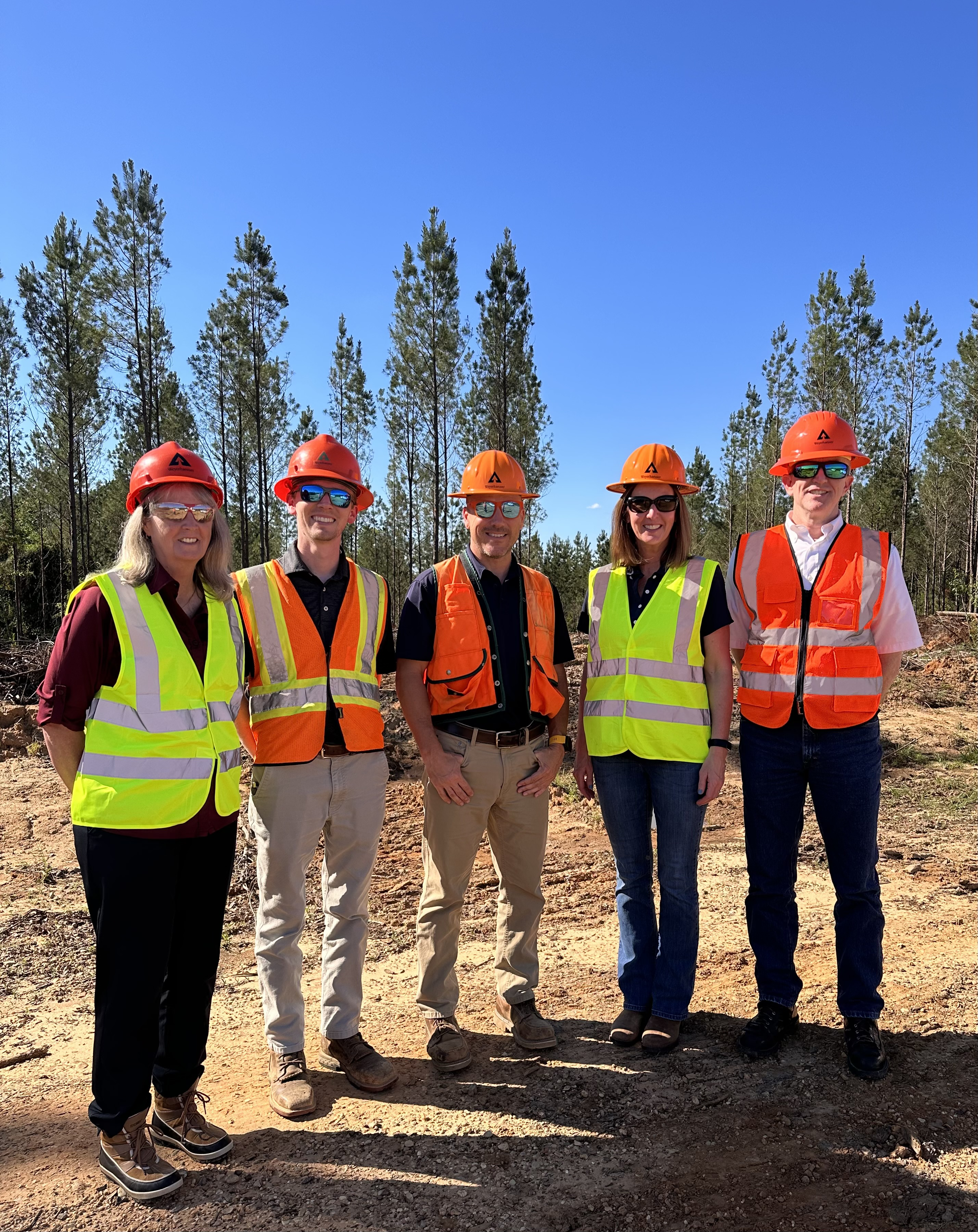 Image of Christie and her team. They are all wearing orange or yellow protective vests and are each wearing hard hats. The day is bright, and all are also wearing sunglasses. In the background trees can be seen.