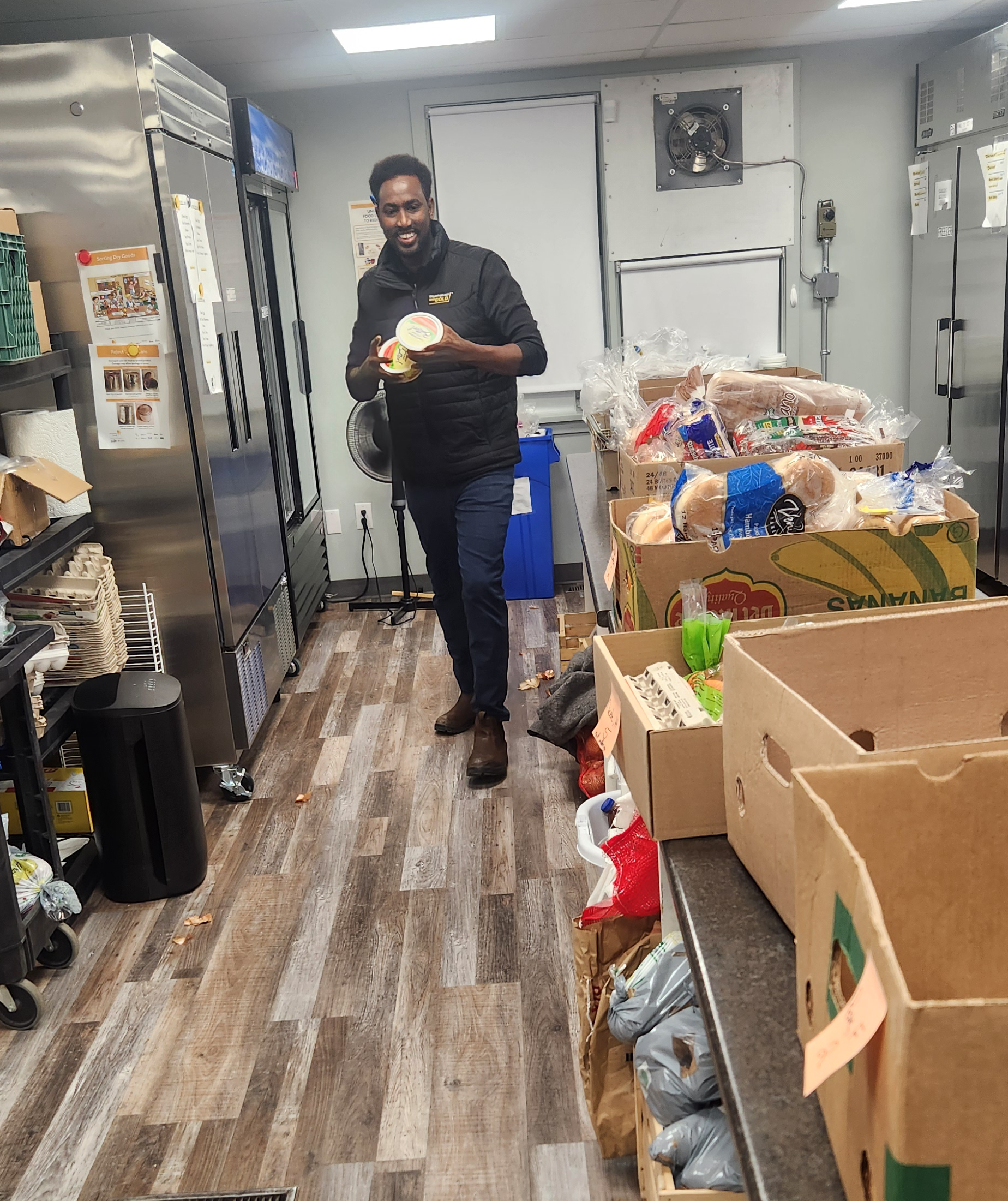 Image of Abdul standing in the Edson Food Bank preparing food. He is in the kitchen and is surrounded by rolls, bread and other supplies.