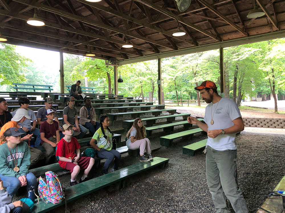 Image of Andrew talking to a group of kids who are sitting on bleachers under a roof at a camp. 