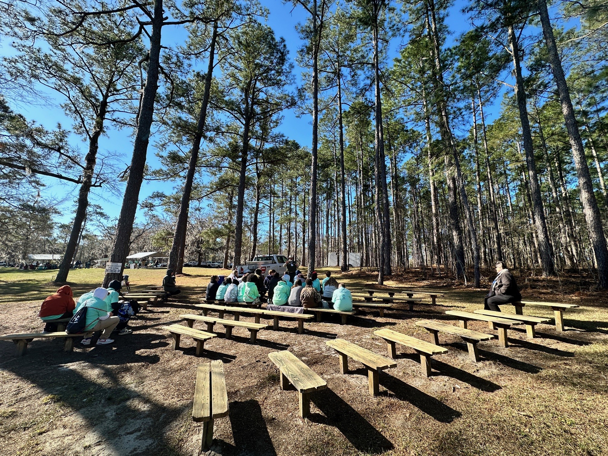 Image of groups of people learning about environmental science during the Envirothon. Benches are lining an area surrounded by trees with groups of poeple wearing turquoise shirts gathered together.