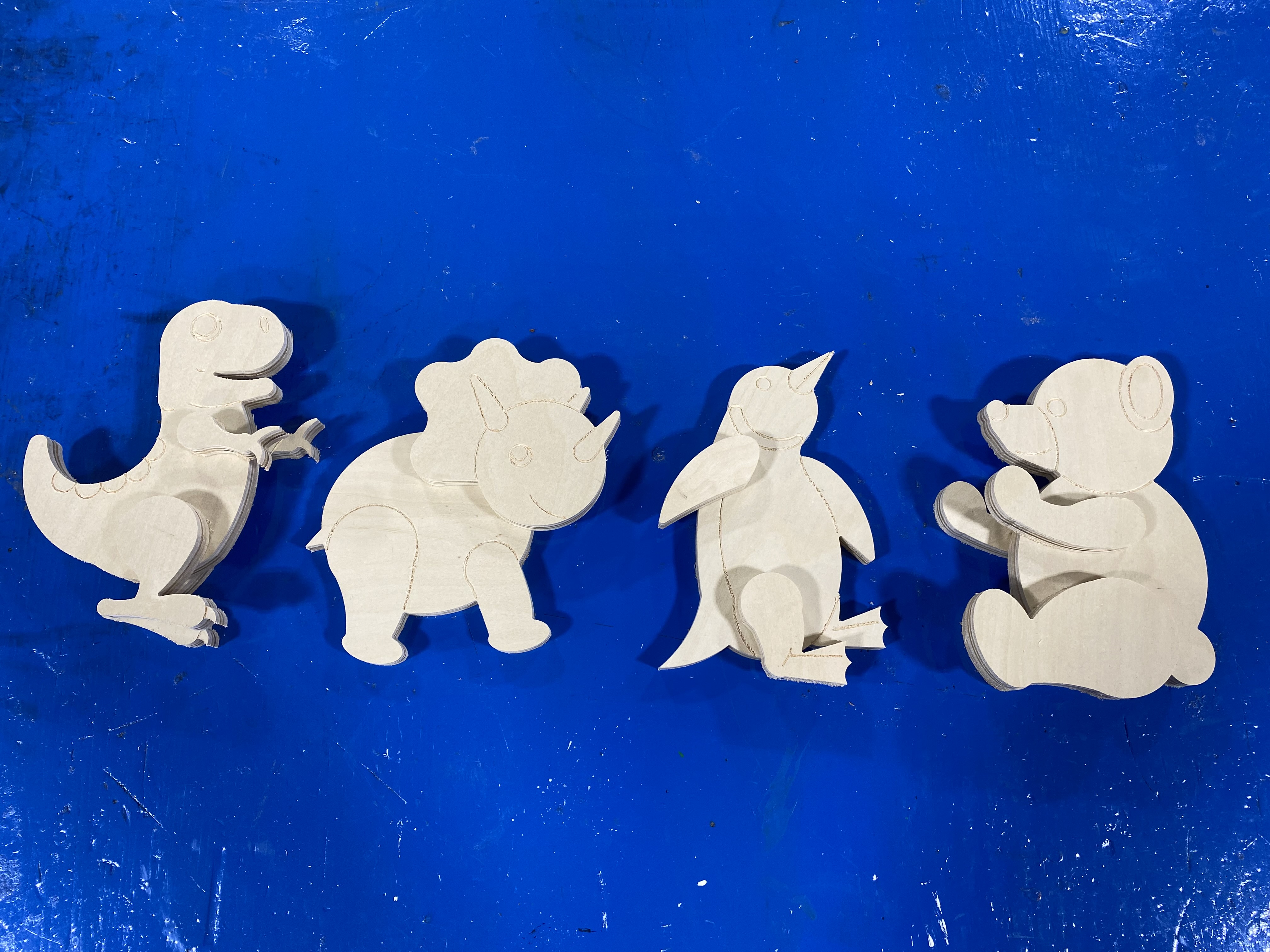 Images of the four wooden toys assembled and ready for painting. The toys are a playful T.Rex, a tiranasaurus, a playful pengui, and a teddy bear.