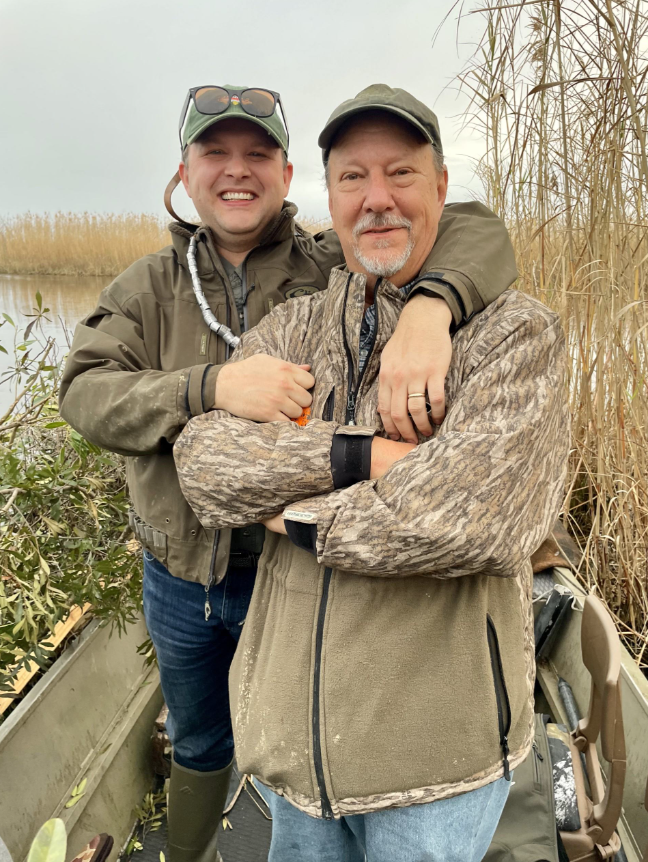 Image of Alex and his friend and former TNC colleague Keith Ouchley. Both are standing in a boat in a marsh and are wearing camouflage jackets.