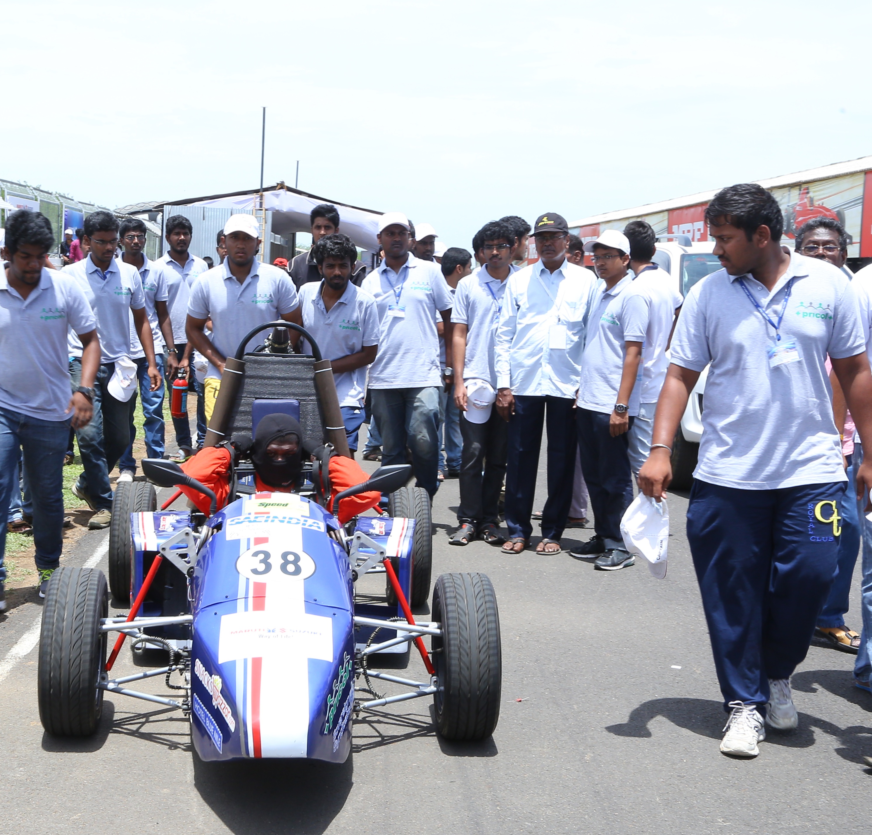 Image showing Satish, standing far right, and his team members from his colleage preparing for an endurance contest. A red, white and blue formula one vehicle is to the left with numerous students standing behind it. 