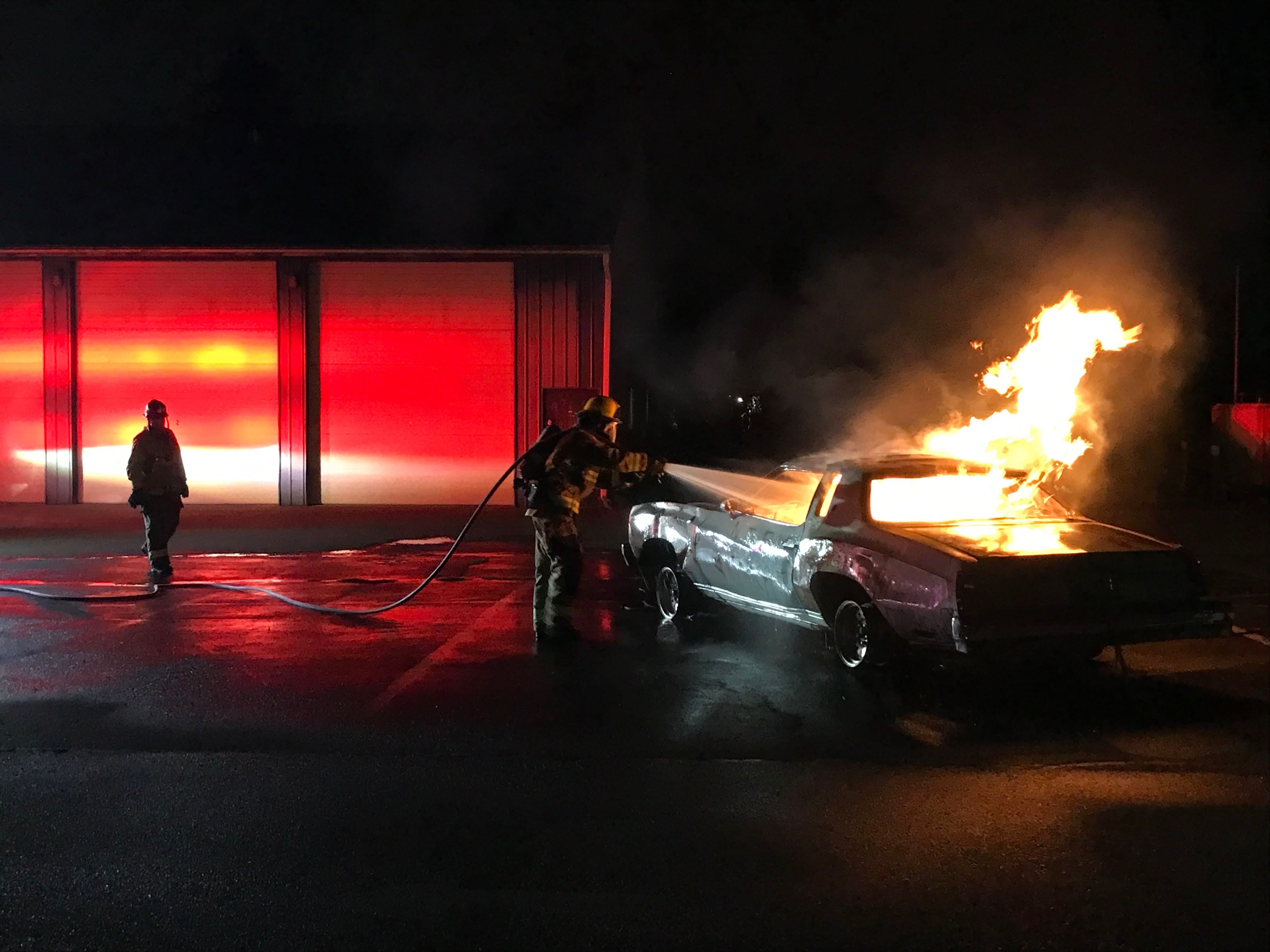 Image of a firefighting drill, which has a car on fire and a fireman approaching spraying water from a hose that is over his shoulder. 