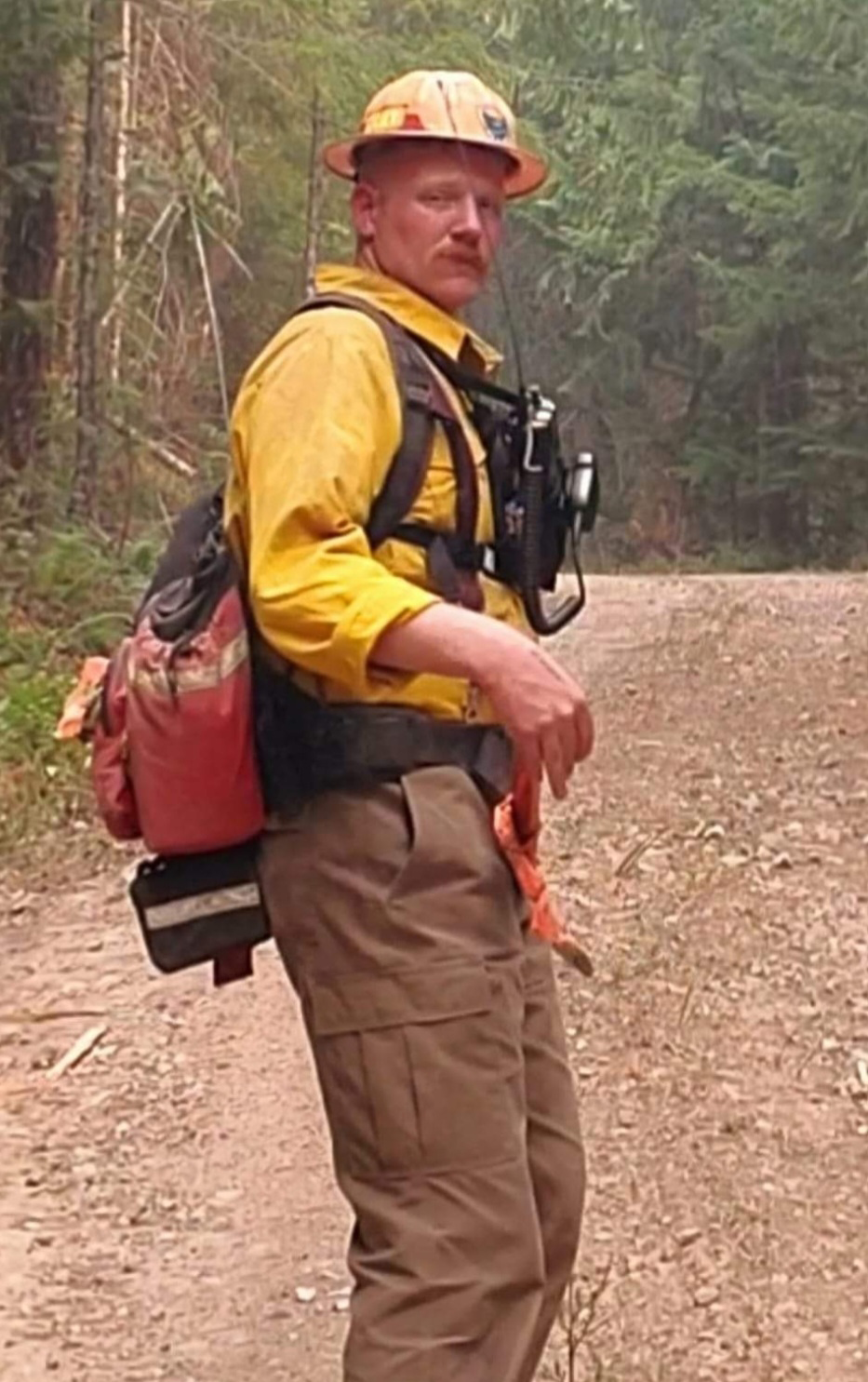 Image of Ruger Sanow, a forest engineer. He's looking back at the camera while wearing a hardhat and yellow shirt and carring firefighting and protective gear. Ruger has a mustache.