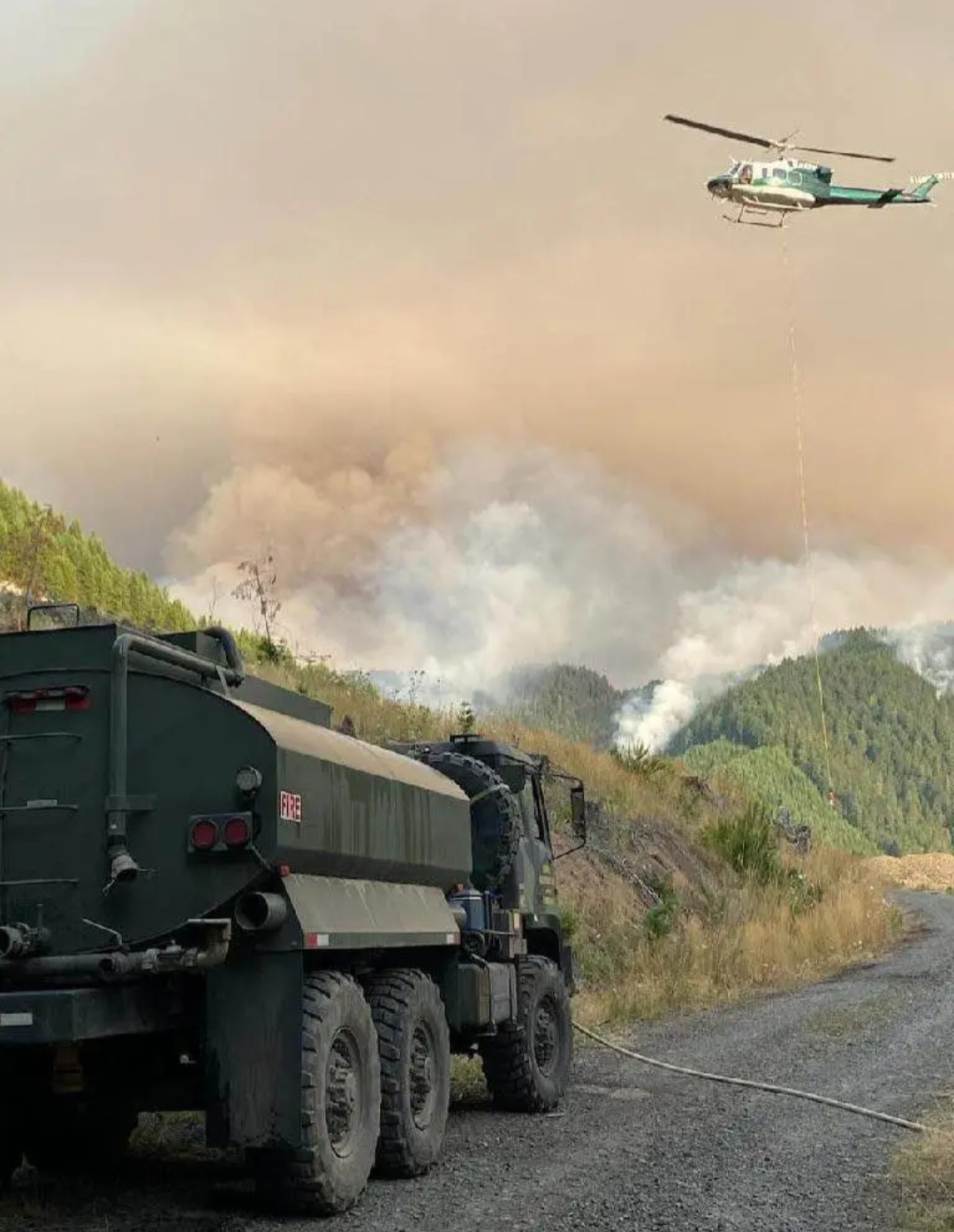 Image of a helicopter and truck with smoke billowing from the forest in hte background.