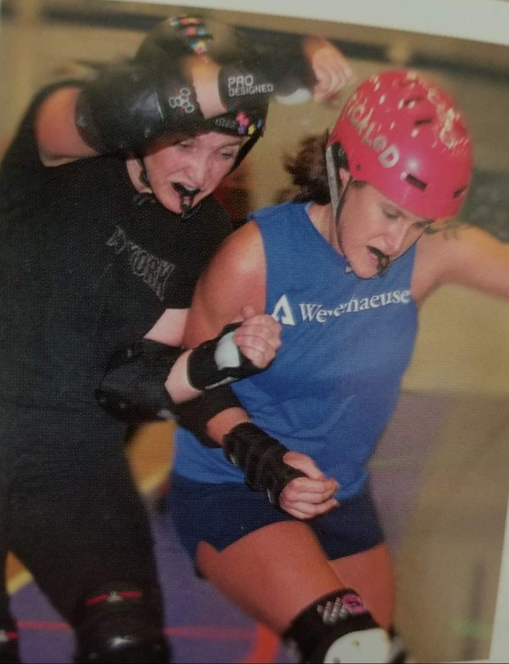 Image of Amber taking part in a roller derby game in 2009. Amber is clad in a red helmet and blue t-shirt with "Weyerhaesuer" on the front, and has an opponent trying to stop her progress.